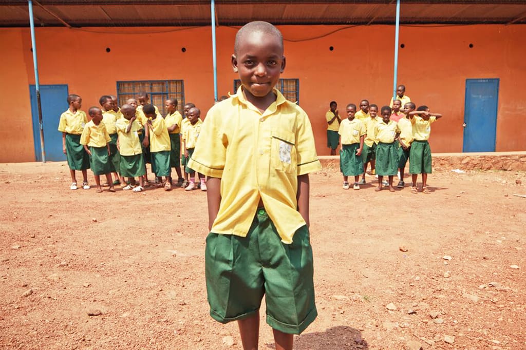 A boy in a uniform stands in front of a group of children with his hands in his pockets and smiles.